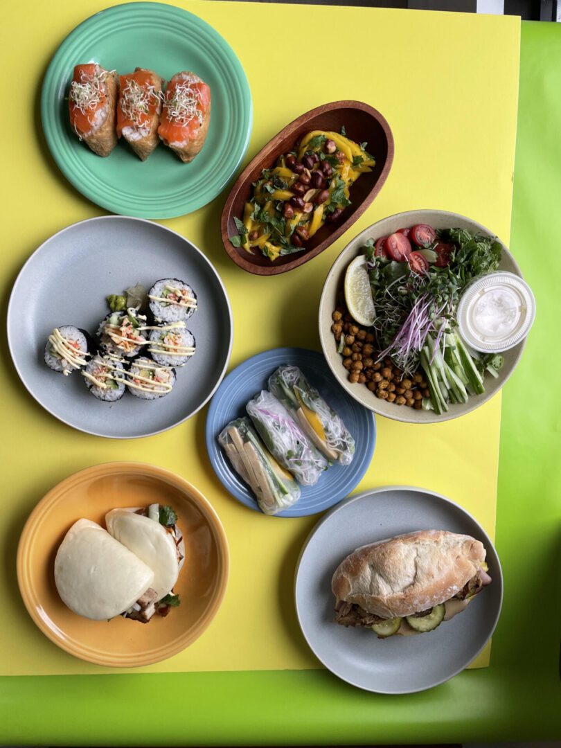 Collection of catering options on a yellow table including sushi, salads, sandwiches and other foods from around the world.